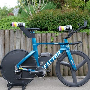 Könnte beinhalten: Ein türkisblaues Factor Hanzo Triathlon-Fahrrad mit schwarzen Rädern und einem schwarzen Sattel. Das Fahrrad hat zwei Wasserflaschen mit bunten Designs. Das Fahrrad steht vor einem Holzzaun und grünem Laub.