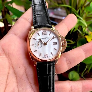 Könnte beinhalten: Eine goldfarbene Panerai Armbanduhr mit weißem Zifferblatt und schwarzem Lederarmband. Die Uhr hat ein quadratisches Zifferblatt mit den Zahlen 12 und 6 sowie ein kleines Zifferblatt auf der 9-Uhr-Position. Die Uhr hat eine Datumsanzeige auf der 3-Uhr-Position.