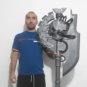 Può includere: Una grande ascia grigia a tema fantasy con un design a teschio e dettagli meccanici. L'ascia ha un lungo manico e una grande lama. La persona nell'immagine sta tenendo l'ascia.