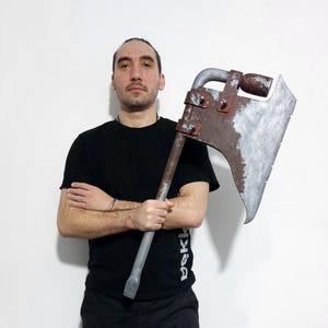 Könnte beinhalten: Eine große, verwitterte Axt mit grauem Stiel und rostiger Metallklinge. Der Axtkopf ist etwa 46 cm breit. Die Axt hat eine gebogene Klinge und ist für den Schwerlastbetrieb ausgelegt. Die Axt ist eine Requisite oder ein Dekorationsartikel.