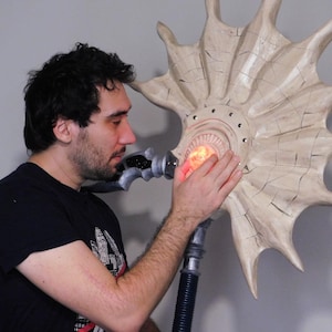 Puede incluir: Una gran escultura en forma de estallido solar estilizado, de color crema, con una luz naranja brillante en el centro. La escultura tiene un mango gris metálico y un tubo negro y acanalado. Una persona está tocando la escultura.