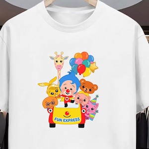 Puede incluir: Camiseta blanca con un dise&ntilde;o de dibujos animados colorido. El gr&aacute;fico muestra un payaso y varios animales de dibujos animados en un veh&iacute;culo amarillo con la inscripci&oacute;n "FUN EXPRESS". El dise&ntilde;o tambi&eacute;n incluye globos y una jirafa.