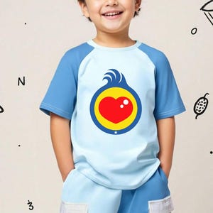 Puede incluir: Camiseta azul claro y azul de manga corta con un gr&aacute;fico en forma de coraz&oacute;n. El gr&aacute;fico presenta un coraz&oacute;n rojo dentro de un c&iacute;rculo amarillo, con olas azules y un punto blanco. La camiseta la lleva un ni&ntilde;o.