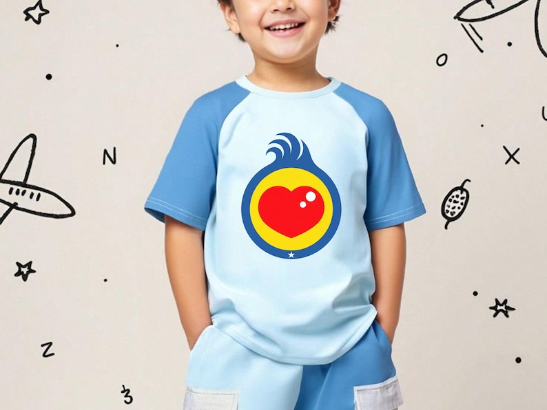 Puede incluir: Camiseta azul claro y azul de manga corta con un gr&aacute;fico en forma de coraz&oacute;n. El gr&aacute;fico presenta un coraz&oacute;n rojo dentro de un c&iacute;rculo amarillo, con olas azules y un punto blanco. La camiseta la lleva un ni&ntilde;o.