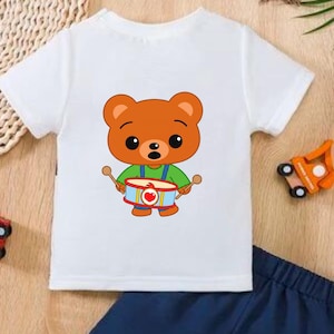 Puede incluir: Camiseta blanca con un oso de dibujos animados tocando un tambor, combinada con pantalones cortos azul marino. El oso es marr&oacute;n con una camisa verde y peto azul. Coches de juguete de madera colocados alrededor de la ropa.