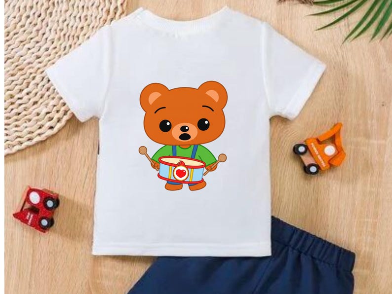 Puede incluir: Camiseta blanca con un oso de dibujos animados tocando un tambor, combinada con pantalones cortos azul marino. El oso es marr&oacute;n con una camisa verde y peto azul. Coches de juguete de madera colocados alrededor de la ropa.