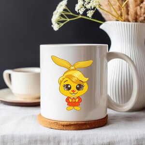 Puede incluir: Taza de cer&aacute;mica blanca con un personaje de dibujos animados con pelo amarillo y un atuendo rojo. La taza est&aacute; sobre un posavasos redondo marr&oacute;n. El fondo incluye una jarra blanca y una taza blanca peque&ntilde;a.