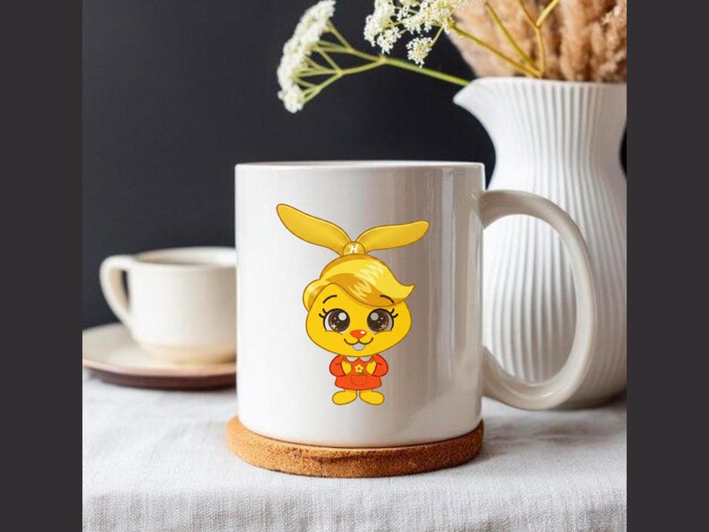 Puede incluir: Taza de cer&aacute;mica blanca con un personaje de dibujos animados con pelo amarillo y un atuendo rojo. La taza est&aacute; sobre un posavasos redondo marr&oacute;n. El fondo incluye una jarra blanca y una taza blanca peque&ntilde;a.
