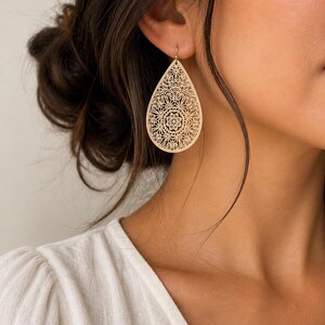 May include: Gold-colored filigree teardrop earrings. The earrings feature an intricate floral design and are suspended from a simple hook. The earrings are a light gold color and are worn on the ear.