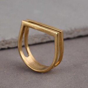 Peut inclure: Une bague dorée au design géométrique. La bague présente une section supérieure rectangulaire et une bande incurvée. La bague est faite d'un matériau métallique brillant. La bague est un bijou.