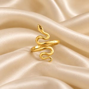 Pode incluir: Um anel de tom dourado em forma de serpente enrolada. A banda de metal forma o corpo da serpente, que se enrola para criar a forma do anel. A cabeça da serpente está posicionada no topo, com um acabamento liso e polido em toda a peça.