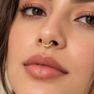 May include: A gold septum ring featuring a twisted rope design with small, clear, round-cut stones embedded along the front surface. The jewelry is circular in shape and sits securely within the nasal septum, showcasing a polished metallic finish.
