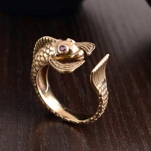 May include: An open-style ring crafted from a metallic gold-toned material, shaped like a fish with detailed scales and fins. The fish features a small, circular, deep pink gemstone set as an eye, with the head and tail ends meeting to form the band.