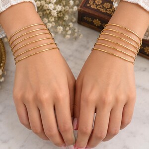 Peut inclure: Un ensemble de douze joncs fins en métal doré poli portés aux poignets. Chaque poignet présente six bracelets circulaires individuels, lisses, uniformes en épaisseur et en couleur, créant un aspect empilé sur les deux bras.