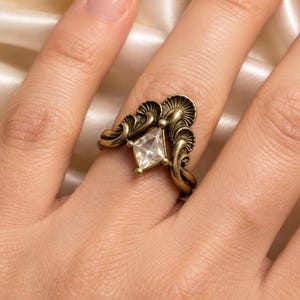 Peut inclure: Une bague en laiton ornée d'une pierre précieuse transparente en forme de cerf-volant au centre. L'anneau présente un travail de métal complexe en forme d'éventail qui s'incurve autour de la pierre, créant une structure symétrique et superposée.