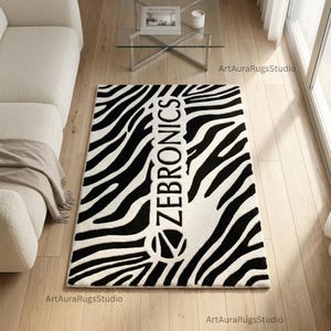 Puede incluir: Una alfombra rectangular con rayas de cebra en blanco y negro. La palabra "ZEBRONICS" está centrada verticalmente en blanco, con una "Z" estilizada en la parte inferior. La alfombra está sobre un suelo de madera clara.