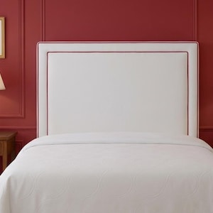 May include: A bedroom with a white bed and patterned duvet, a white headboard with red trim, and two bedside tables with lamps. The walls are painted a deep red. The lamps have beige shades and the artwork is framed.