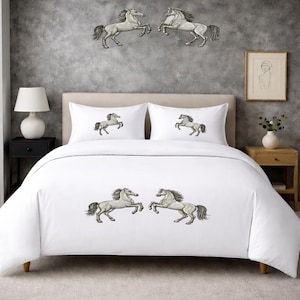 Könnte beinhalten: Weißes Bettwäsche-Set mit gestickten Pferdemotiven. Das Set enthält einen Bettbezug und zwei Kissenbezüge mit passenden Pferdekunstwerken. An der Wand hinter dem Bett befinden sich zwei Metallpferd-Skulpturen.