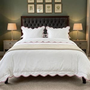 May include: A white bedspread with a red scalloped edge, paired with matching pillowcases. The bed is made up with a beige throw blanket and set against a dark gray tufted headboard. Two bedside lamps flank the bed.