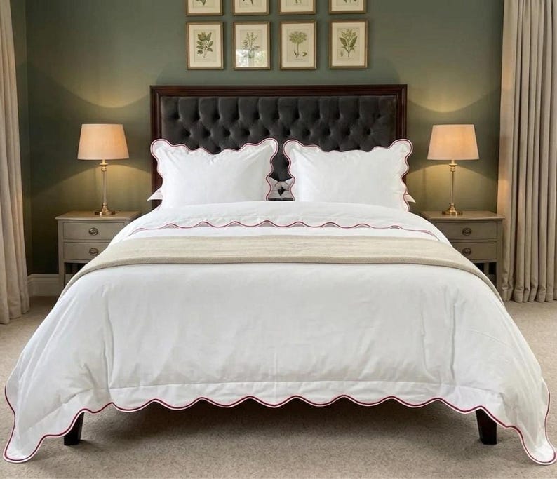 May include: A white bedspread with a red scalloped edge, paired with matching pillowcases. The bed is made up with a beige throw blanket and set against a dark gray tufted headboard. Two bedside lamps flank the bed.