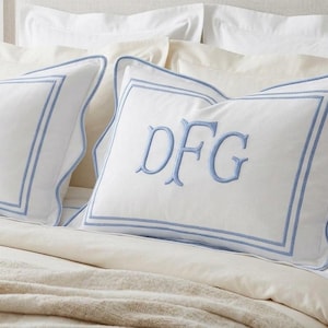 May include: Two white rectangular pillow shams featuring a scalloped edge and a double-line blue border. Each sham is embroidered with the blue monogram letters DFG in a serif font. The pillows are arranged on a bed with cream-colored bedding.