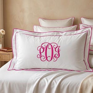 May include: White pillowcase with a magenta embroidered monogram and trim. The bedding is on a white bed with a neutral-toned background. The pillow has a decorative border.