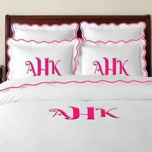 May include: A white bedding set featuring four pillow shams and a duvet cover, all accented with a scalloped pink trim. Each piece is embroidered with the monogram AHK in a bold, bright pink font, centered on the white fabric surface.
