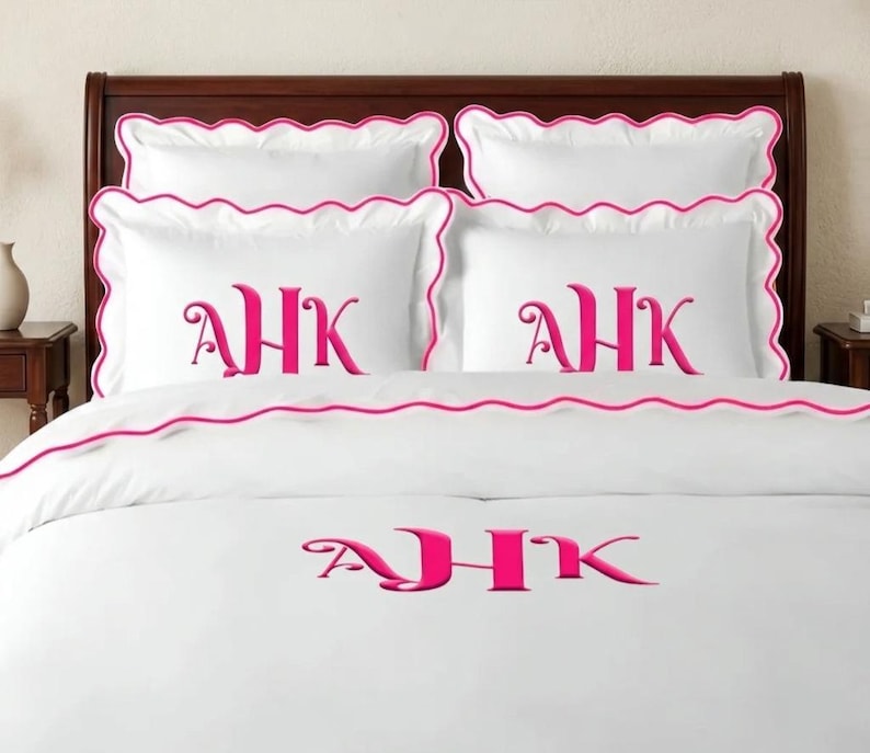 May include: A white bedding set featuring four pillow shams and a duvet cover, all accented with a scalloped pink trim. Each piece is embroidered with the monogram AHK in a bold, bright pink font, centered on the white fabric surface.