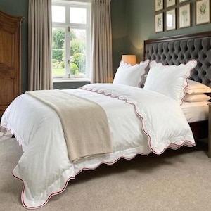 May include: A white bedspread and pillow set with a red scalloped trim. A beige throw blanket is draped across the bed. The bed is in a room with a dark gray upholstered headboard, a window, and a wooden armoire.