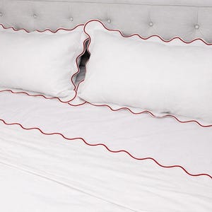 May include: White bedding set with red scalloped trim. The set includes two pillowcases and a fitted sheet. The bedding is on a bed with a gray upholstered headboard. The sheets and pillowcases are made of a soft, smooth fabric.