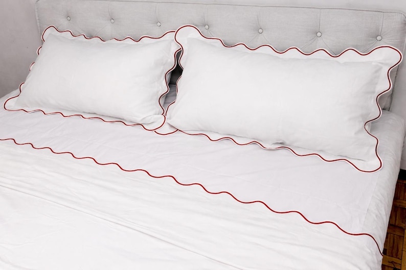 May include: White bedding set with red scalloped trim. The set includes two pillowcases and a fitted sheet. The bedding is on a bed with a gray upholstered headboard. The sheets and pillowcases are made of a soft, smooth fabric.