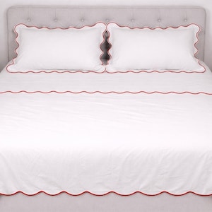 May include: White bedding set with red scalloped trim. The set includes two pillowcases and a fitted sheet, all with a decorative red border. The bed is made and ready for use.