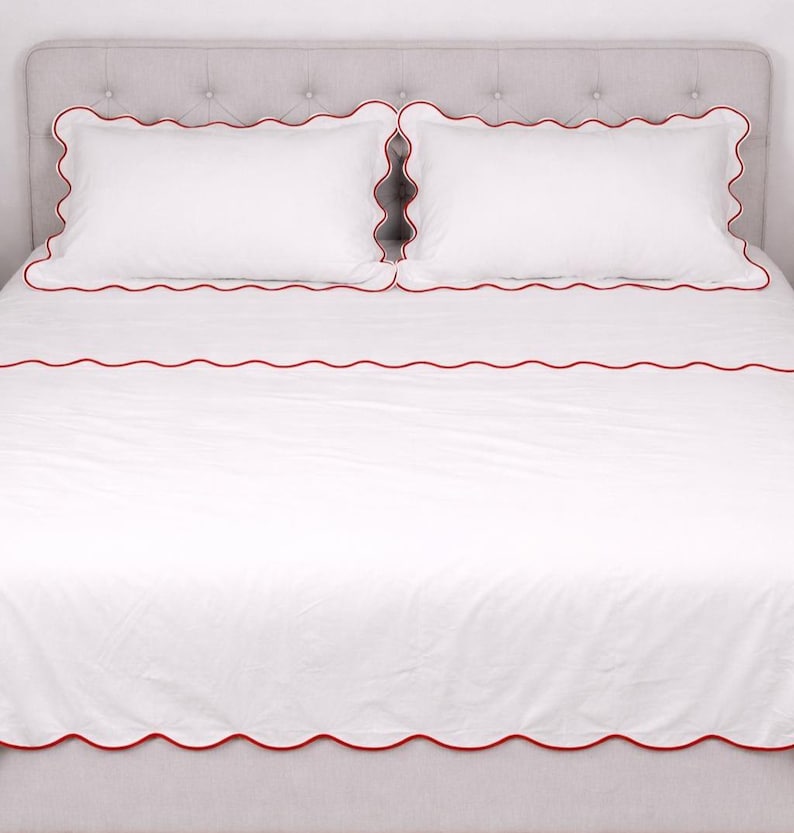 May include: White bedding set with red scalloped trim. The set includes two pillowcases and a fitted sheet, all with a decorative red border. The bed is made and ready for use.