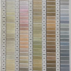 May include: A color chart featuring five vertical columns of thread samples arranged in rows. Each row displays a specific thread color alongside its corresponding alphanumeric code. The palette includes shades of white, cream, pink, green, gold, and blue.