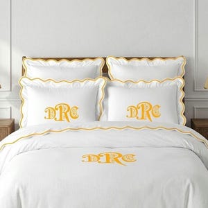 May include: A white bedding set featuring a scalloped edge with yellow trim. The set includes two standard pillows and two shams, each embroidered with a yellow monogram reading "DRC". A matching white duvet cover also displays the yellow "DRC" monogram.
