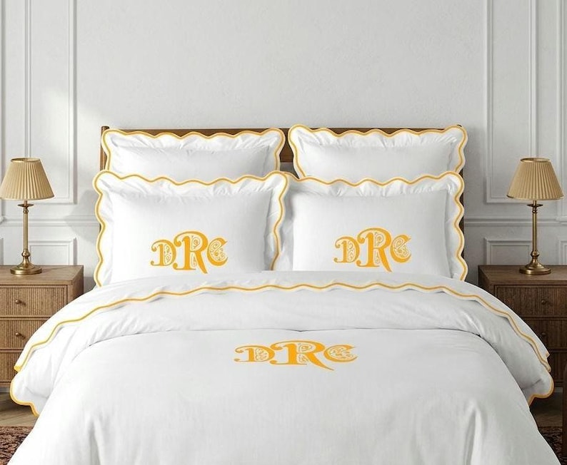 May include: A white bedding set featuring a scalloped edge with yellow trim. The set includes two standard pillows and two shams, each embroidered with a yellow monogram reading "DRC". A matching white duvet cover also displays the yellow "DRC" monogram.
