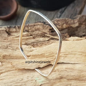 May include: A silver-colored, open-ended bracelet with a geometric, angular design. The bracelet is a continuous, slightly flattened oval shape. The background is a textured, light brown wooden surface. The text "arpitshinejewels" is visible.