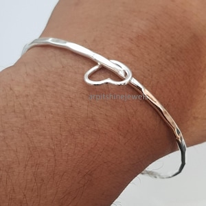May include: A silver-colored bangle bracelet with a hammered texture and a heart-shaped knot design. The bracelet is a simple, elegant piece of jewelry.