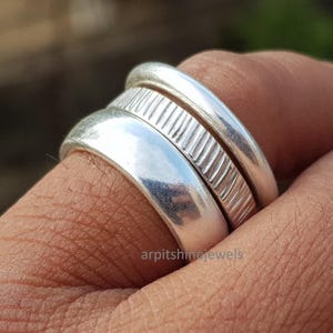 May include: Close-up of three silver rings stacked on a finger. The rings have different designs, including a smooth band, a textured band, and a rounded band. The rings are made of a shiny, reflective metal.