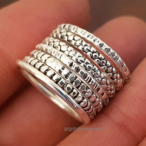 May include: A stackable silver ring set with seven bands. Each band has a unique texture, including dotted, ridged, and smooth designs. The rings are displayed on a hand, showcasing the intricate details and the overall style of the jewelry.