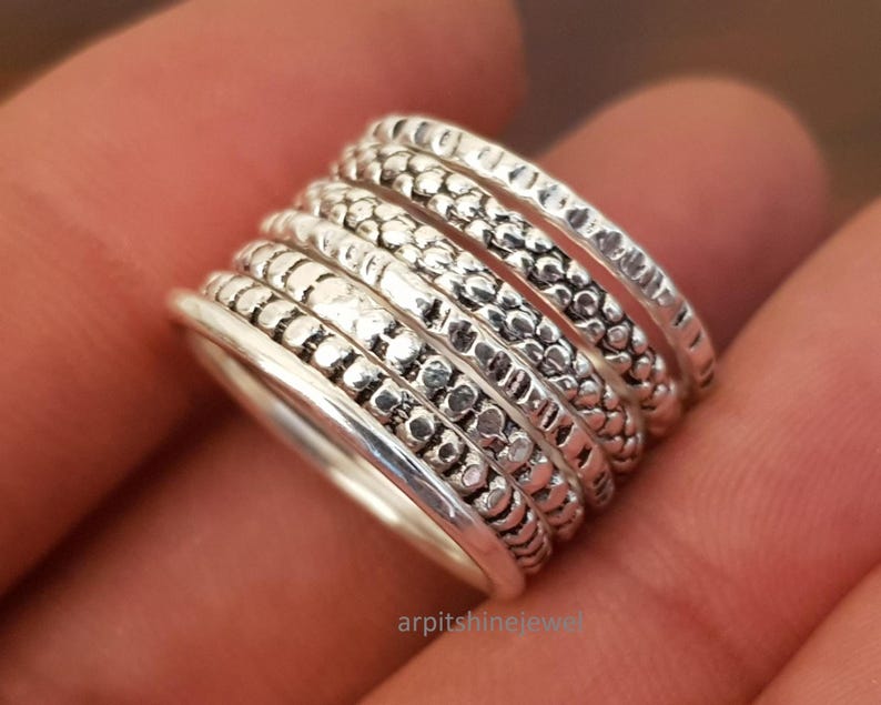 May include: A stackable silver ring set with seven bands. Each band has a unique texture, including dotted, ridged, and smooth designs. The rings are displayed on a hand, showcasing the intricate details and the overall style of the jewelry.