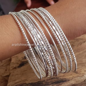 May include: A selection of silver bangles, featuring diverse textures such as twisted rope and hammered patterns. The bangles are displayed on a wrist, highlighting their varied designs and reflective qualities. The backdrop is a wooden surface.