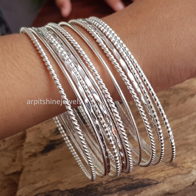 May include: A selection of silver bangles, featuring diverse textures such as twisted rope and hammered patterns. The bangles are displayed on a wrist, highlighting their varied designs and reflective qualities. The backdrop is a wooden surface.