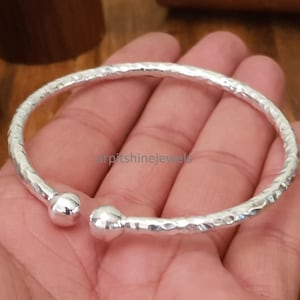 Op de afbeelding: Een zilverkleurige, gehamerde manchetarmband met afgeronde kogeluiteinden. Het open ontwerp maakt het gemakkelijk om te dragen. De armband wordt op een hand getoond, wat de grootte en het ontwerp benadrukt. Het metaal heeft een heldere, reflecterende kwaliteit.