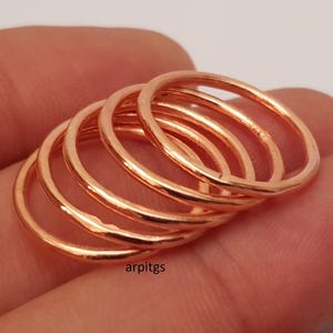 May include: A stack of five thin, rose gold rings. The rings are smooth and shiny, with a simple, minimalist design. The rings are stacked together, creating a layered effect. The rings are likely made of metal.