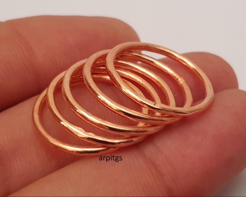May include: A stack of five thin, rose gold rings. The rings are smooth and shiny, with a simple, minimalist design. The rings are stacked together, creating a layered effect. The rings are likely made of metal.