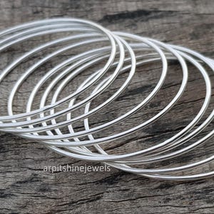 May include: A stack of silver bangles, each with a simple, rounded design. The bracelets are arranged on a weathered wooden surface, creating a contrast between the shiny metal and the rustic background. The jewelry is elegant and minimalist.
