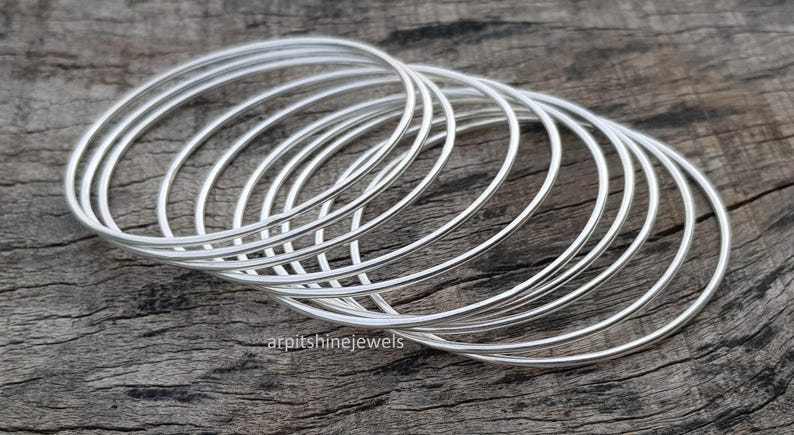 May include: A stack of silver bangles, each with a simple, rounded design. The bracelets are arranged on a weathered wooden surface, creating a contrast between the shiny metal and the rustic background. The jewelry is elegant and minimalist.