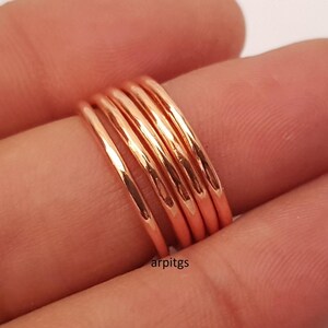 May include: A stack of five thin, rose gold rings. The rings are smooth and shiny, reflecting light. The rings are stacked on a finger, with the skin tone of the finger visible in the background. The rings are a simple, elegant design.