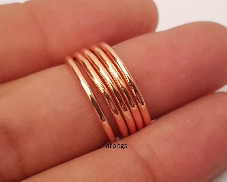 May include: A stack of five thin, rose gold rings. The rings are smooth and shiny, reflecting light. The rings are stacked on a finger, with the skin tone of the finger visible in the background. The rings are a simple, elegant design.